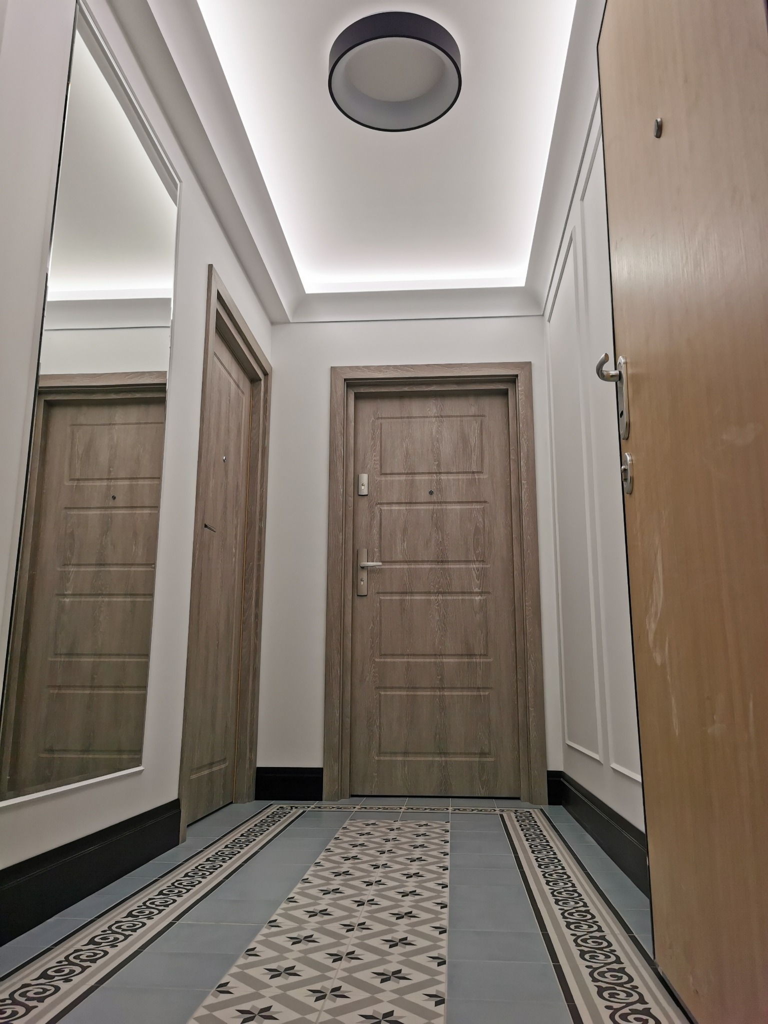 Nowoczesny korytarz z jasnymi ścianami, ozdobnymi listwami, lustrem, wzorzystą podłogą z płytek i drzwiami w odcieniach szarości i drewna, oświetlony ledowym światłem w suficie.