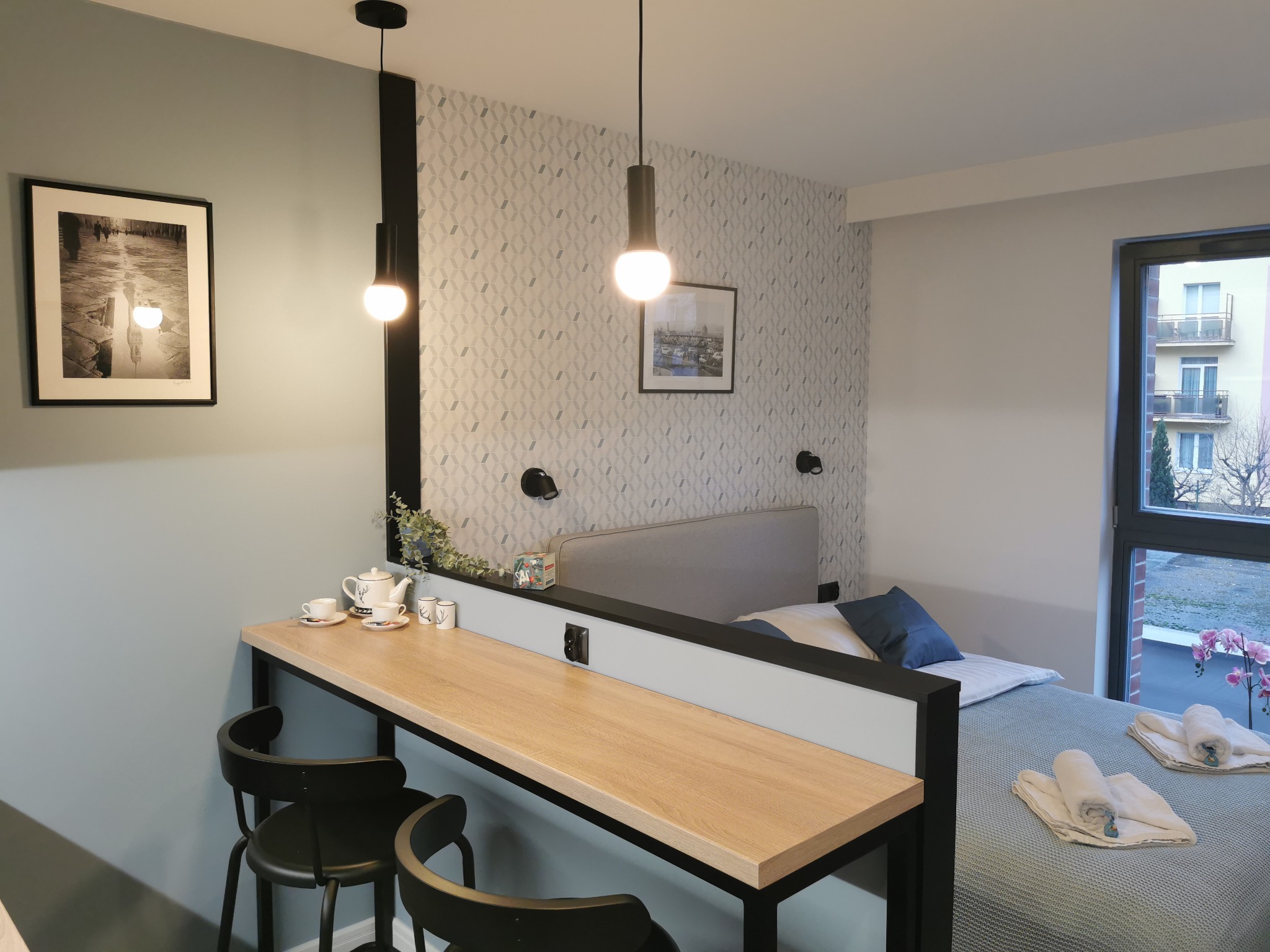 Nowocześnie urządzone studio z aneksem kuchennym oddzielonym od części sypialnej blatem barowym z krzesłami, oświetlonym wiszącymi lampami. Na ścianie dekoracyjna tapeta i obrazy.