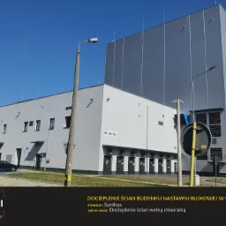 Szary budynek przemysłowy z docieplonymi ścianami, widoczne elementy instalacji i zaparkowane samochody, błękitne niebo w tle.