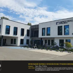 Wykończona elewacja budynku biurowego z dekoracyjnym tynkiem imitującym beton, czarne obramowania okien, logo firmy Spec Bau Polska na fasadzie, zagospodarowany teren zielenią i kamieniami.