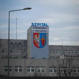 Odnowiona ściana szczytowa Powiatowego Szpitala w Chrzanowie z herbem powiatu, widoczna z poziomu ulicy, oświetlona latarnią uliczną, na tle pochmurnego nieba.