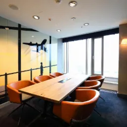Sala konferencyjna z długim drewnianym stołem, pomarańczowymi krzesłami i dekoracyjną folią na szklanych ścianach z motywem lotniczym i sylwetką człowieka.