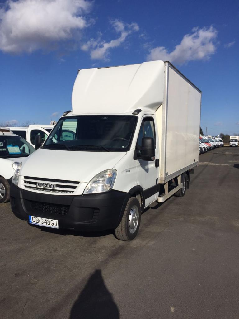 Biały samochód dostawczy Iveco z wysoką zabudową kontenerową, zaparkowany na parkingu z rzędem innych pojazdów w tle pod błękitnym niebem z chmurami.