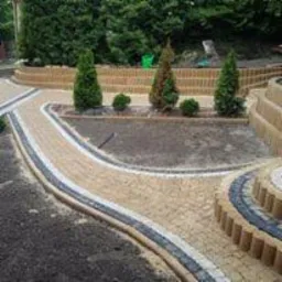 Kaskadowy ogród z ścieżkami z beżowej kostki brukowej obrzeżonej ciemnoszarą kostką, rabaty z niskimi iglakami, w tle zielona roślinność.