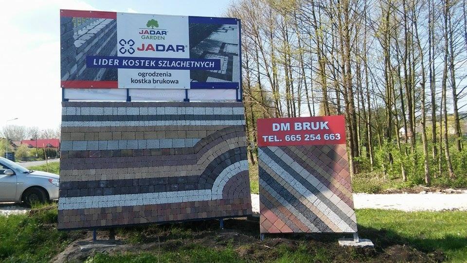 Ekspozycja różnych wzorów i kolorów kostki brukowej na dwóch tablicach reklamowych z logo producenta Jadar Garden i firmy DM Bruk z numerem telefonu, umieszczonych na trawniku.