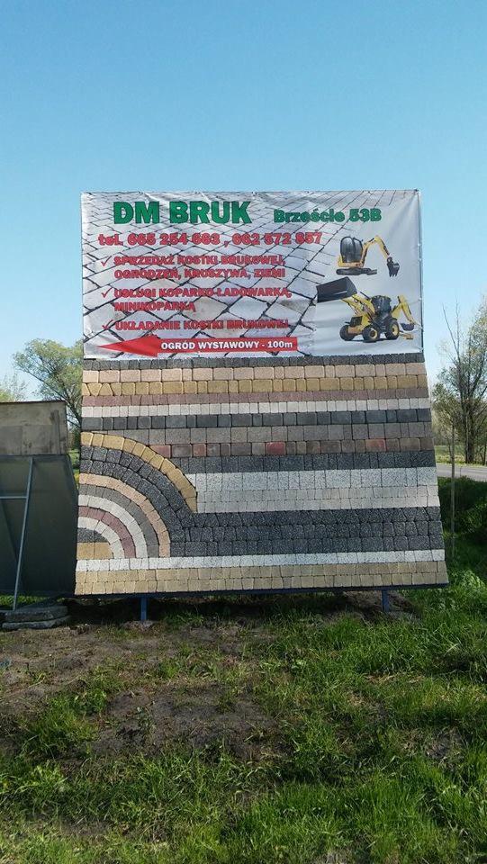 Ekspozycja różnych wzorów i kolorów kostki brukowej ułożonych w poziome pasy i łuki, z widoczną reklamą firmy DM BRUK w tle, informującą o sprzedaży kostki, ogrodzeń, kruszywa i usługach...