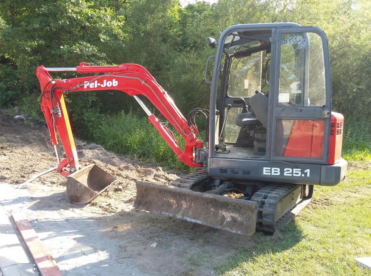 Czerwona mini koparka Pel-Job EB 25.1 stoi na częściowo wyrównanym terenie z widocznymi śladami pracy, z zielenią w tle.