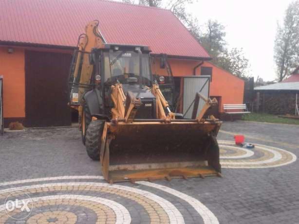 Żółto-czarna koparko-ładowarka Case na brukowanym podwórku z pomarańczowym budynkiem w tle, widoczne ślady użytkowania na łyżce.