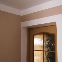 Ściana w odcieniach beżu z białą listwą przysufitową i obramowaniem drzwi z szybą ornamentową.