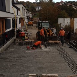 Rolka, Wykopy, Przyłącza, Transport - Dwóch pracowników w pomarańczowych koszulkach układa szarą kostkę brukową na podjeździe przed szeregiem nowoczesnych domów, z koparką i mini-koparką w tle. Widoczne narzędzia i palety z kostką.