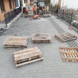 Rolka, Wykopy, Przyłącza, Transport - Dwóch pracowników w pomarańczowych koszulkach układa szarą kostkę brukową na podjeździe przed nowymi domami, widoczne palety z kostką, maszyny budowlane i narzędzia.