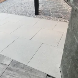 Jasnoszare, prostokątne płyty chodnikowe ułożone w Jamnicy, obok ścieżki z drobnego, białego żwiru i kamiennych płyt, z fragmentem ściany budynku w tle.