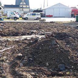 Rolka, Wykopy, Przyłącza, Transport - Przygotowany teren pod budowę z widocznymi betonowymi słupkami, ciężki sprzęt budowlany w tle, w tym wywrotka i ładowarka.