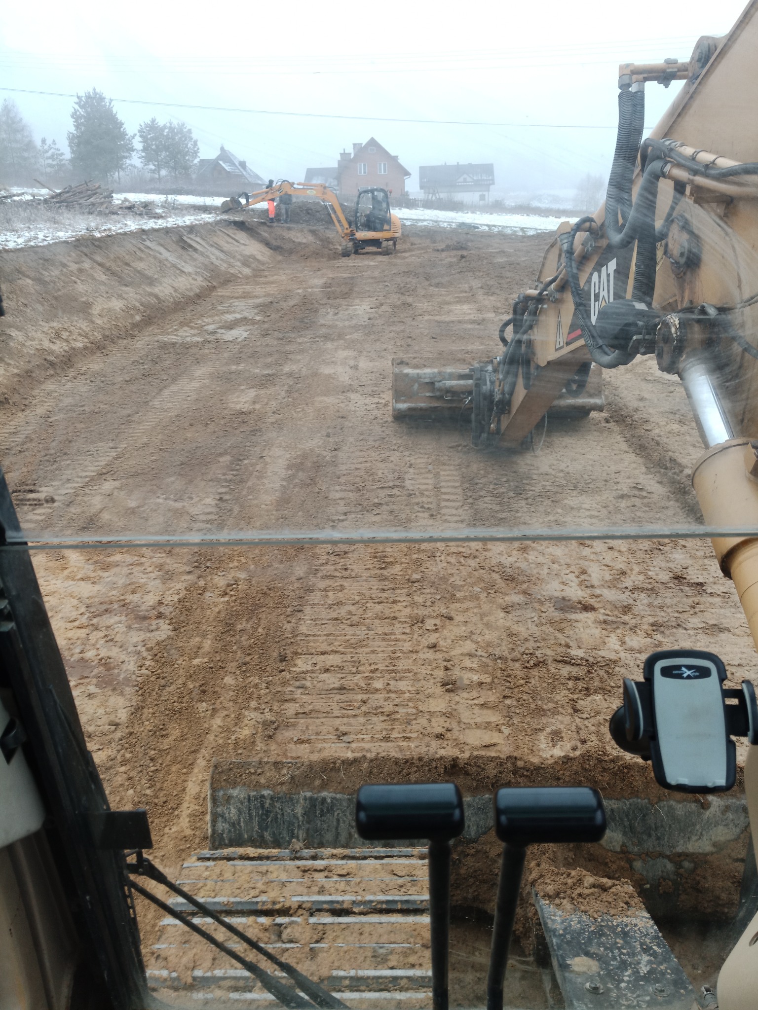 Wykopany rów w ziemi widziany z kabiny koparki CAT, w tle pracująca koparka i operator w pomarańczowym ubraniu, lekko ośnieżony teren, mglista pogoda.