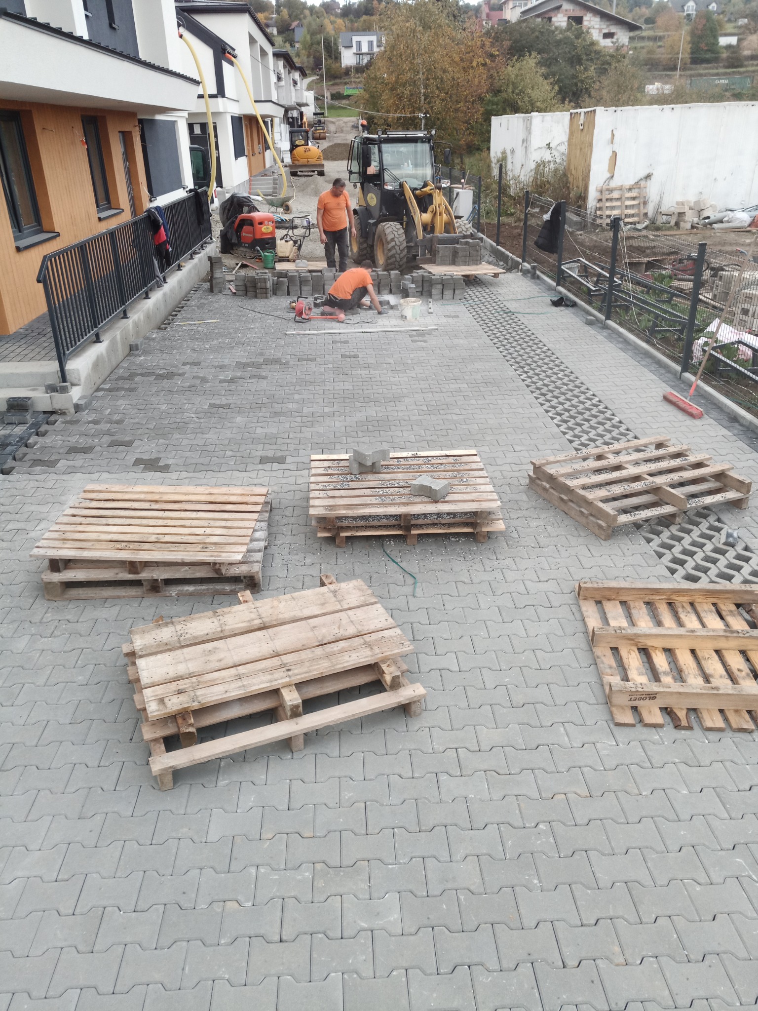 Dwóch pracowników w pomarańczowych koszulkach układa szarą kostkę brukową na podjeździe przed nowymi domami, widoczne palety z kostką, maszyny budowlane i narzędzia.
