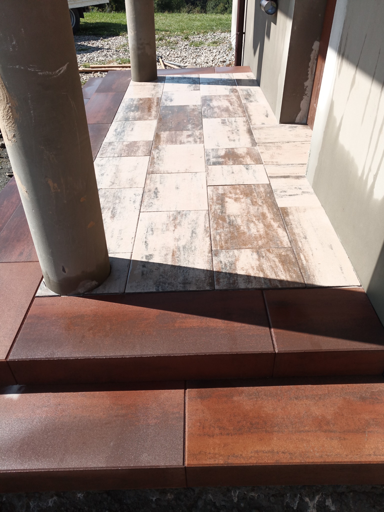 Wykończone wejście do budynku z dwoma stopniami z brązowej kostki i jasną, wzorzystą nawierzchnią, kolumny podtrzymujące zadaszenie.