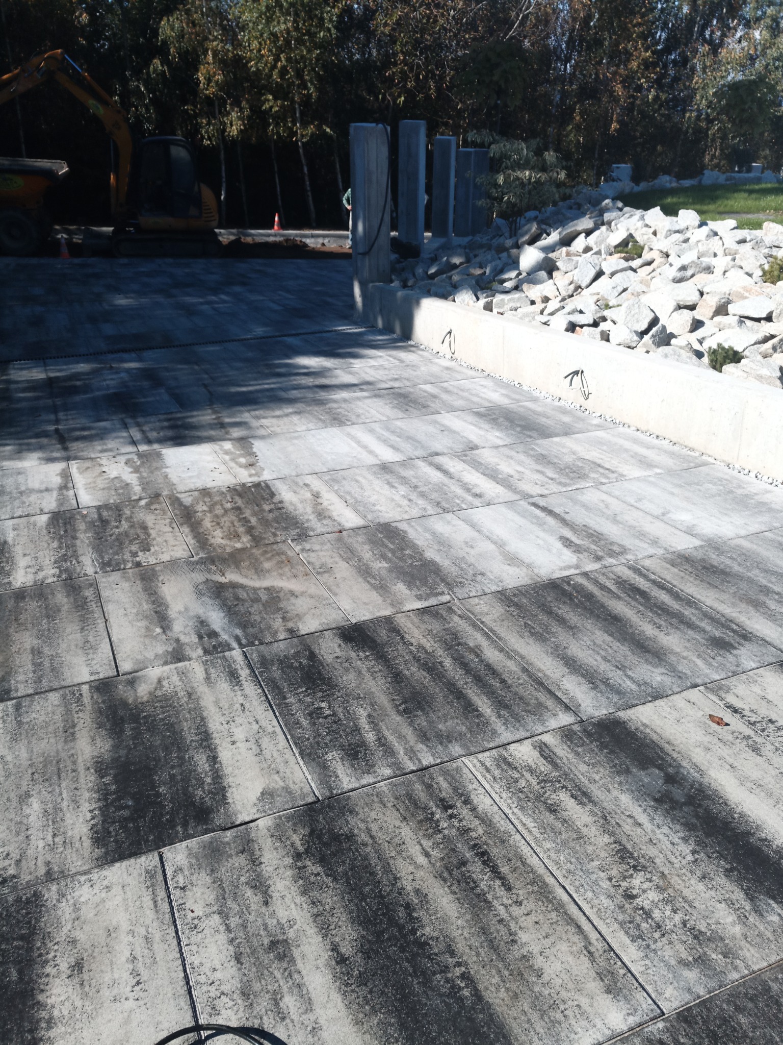 Ułożona szara kostka brukowa z widocznym drenażem, w tle koparka i elementy betonowe oraz nasyp z białych kamieni.