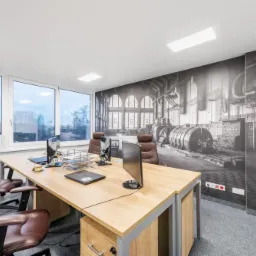 Jasne biuro z drewnianym biurkiem, skórzanymi krzesłami i fototapetą przedstawiającą wnętrze hali fabrycznej.