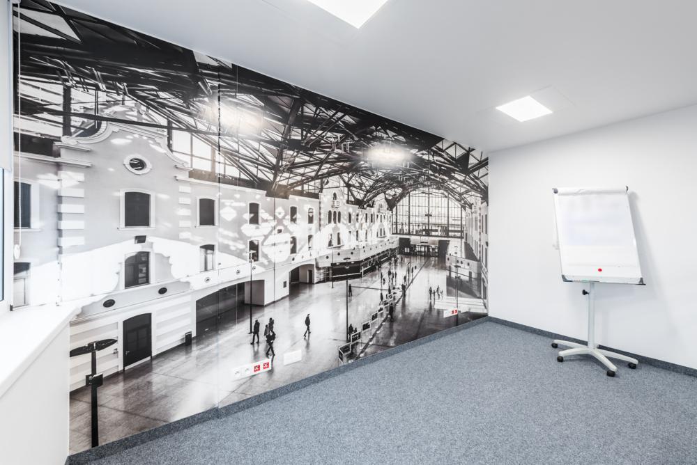 Białe biuro z fototapetą przedstawiającą czarno-biały obraz hali dworca kolejowego z ludźmi, z flipchartem po prawej stronie i szarą wykładziną.