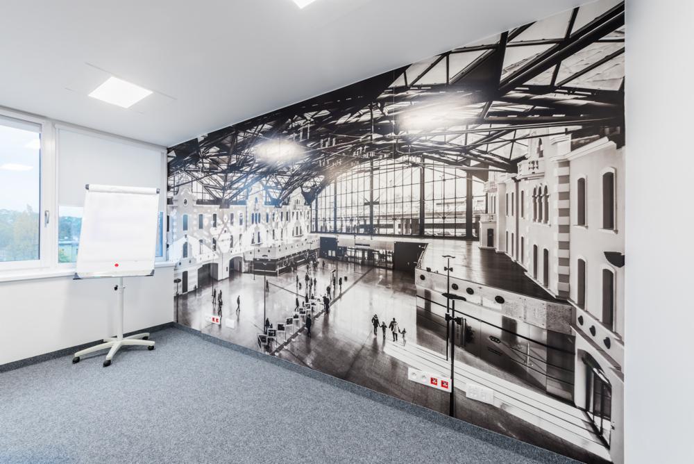 Białe biuro z szarą wykładziną i fototapetą przedstawiającą czarno-biały obraz dworca kolejowego z wysokim, przeszklonym dachem.