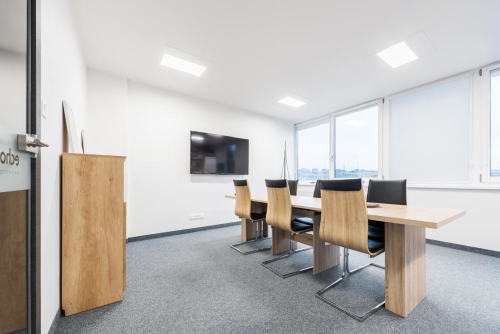 Jasne biuro z drewnianym stołem konferencyjnym, krzesłami z czarnymi siedziskami i dużym telewizorem na ścianie