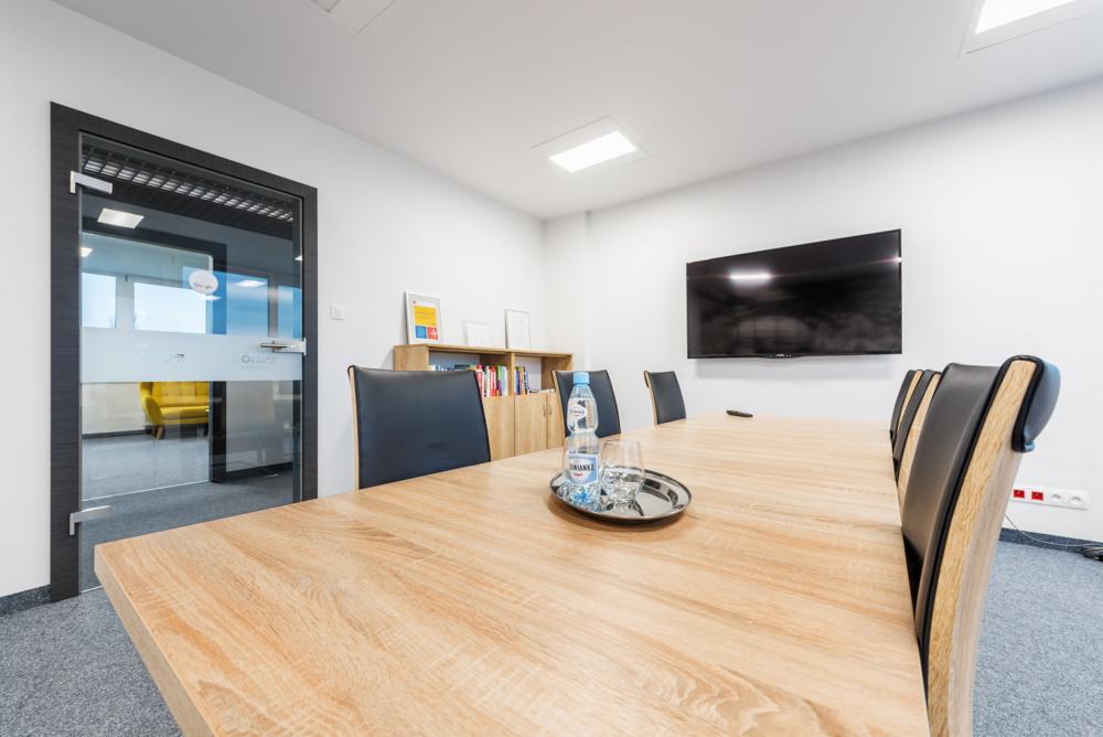 Sala konferencyjna z dużym drewnianym stołem, czarnymi krzesłami, telewizorem na ścianie i szklanymi drzwiami z widoczną żółtą sofą w tle.