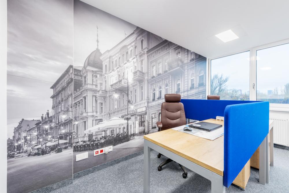 Biuro z fototapetą przedstawiającą starą zabudowę miejską, brązowe krzesło biurowe, biurko z niebieskimi przegrodami i laptopem.