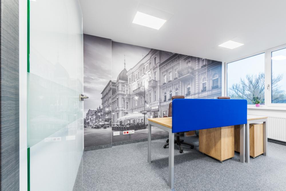 Nowoczesne biuro z fototapetą przedstawiającą starą zabudowę miejską, biurka z niebieskimi przegrodami i przeszklone drzwi z zielonym akcentem.