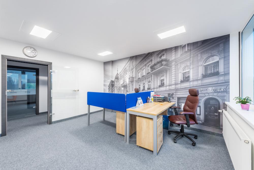 Nowoczesne biuro z biurkami oddzielonymi niebieskimi przegrodami, fototapetą z motywem kamienicy i szarym dywanem.