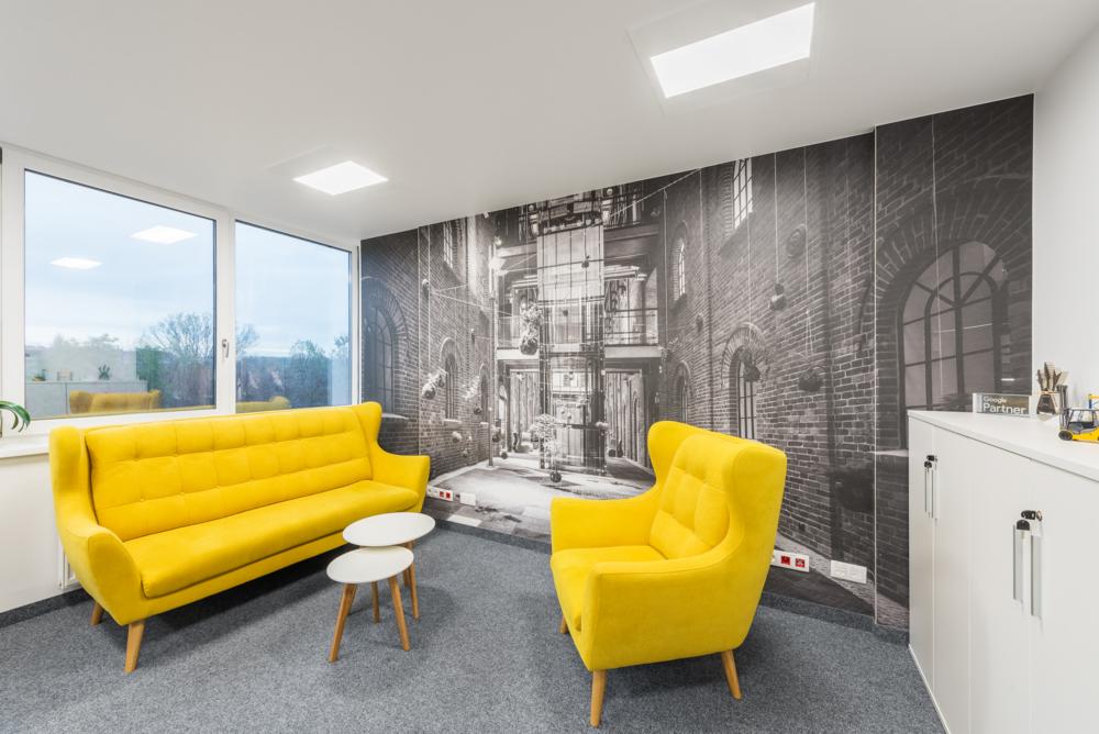 Jasne biuro z żółtą sofą i fotelem na tle fototapety z motywem starej architektury miejskiej.