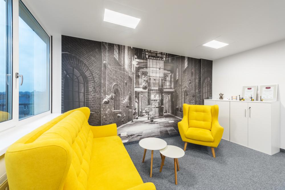 Jasnożółty fotel i sofa w biurze z fototapetą przedstawiającą uliczkę miejską, dwie białe okrągłe stoliki kawowe, biała szafka z certyfikatami.