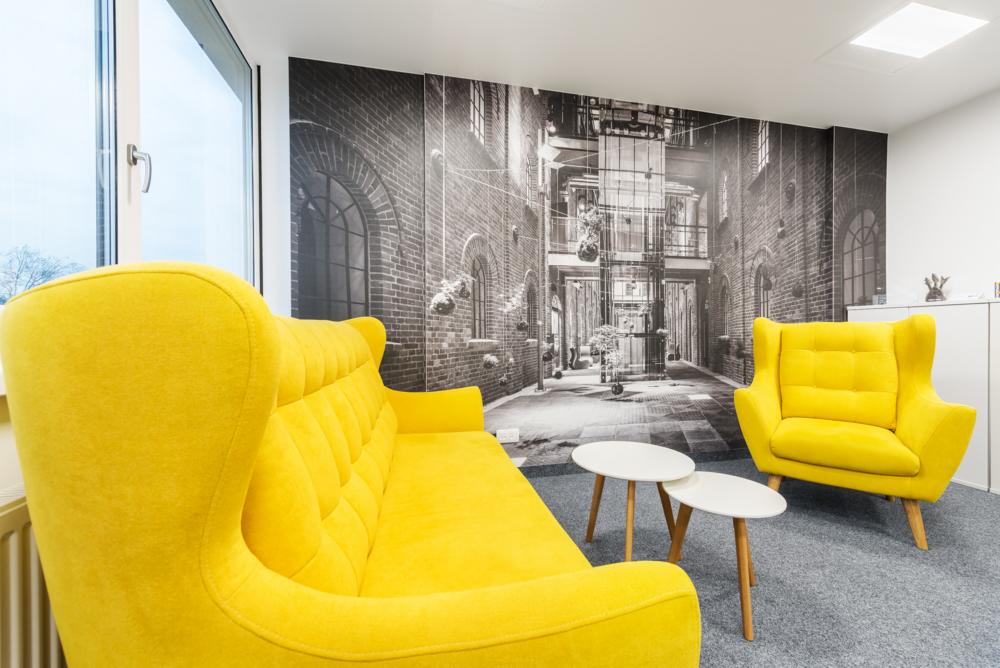 Żółty fotel i sofa w biurze z fototapetą przedstawiającą uliczkę.