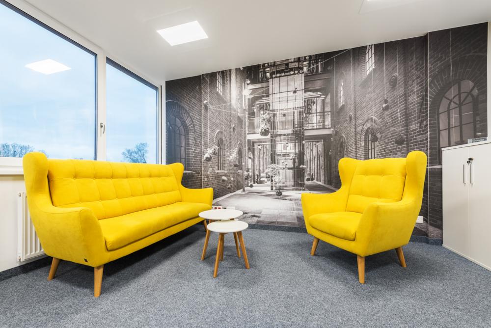 Żółty fotel i sofa w biurze z fototapetą przedstawiającą uliczkę miejską.