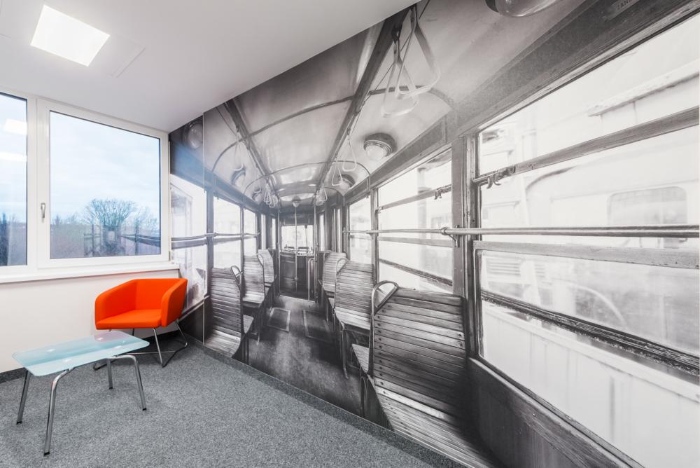 Biuro z fototapetą przedstawiającą wnętrze tramwaju, pomarańczowy fotel i szklany stolik.