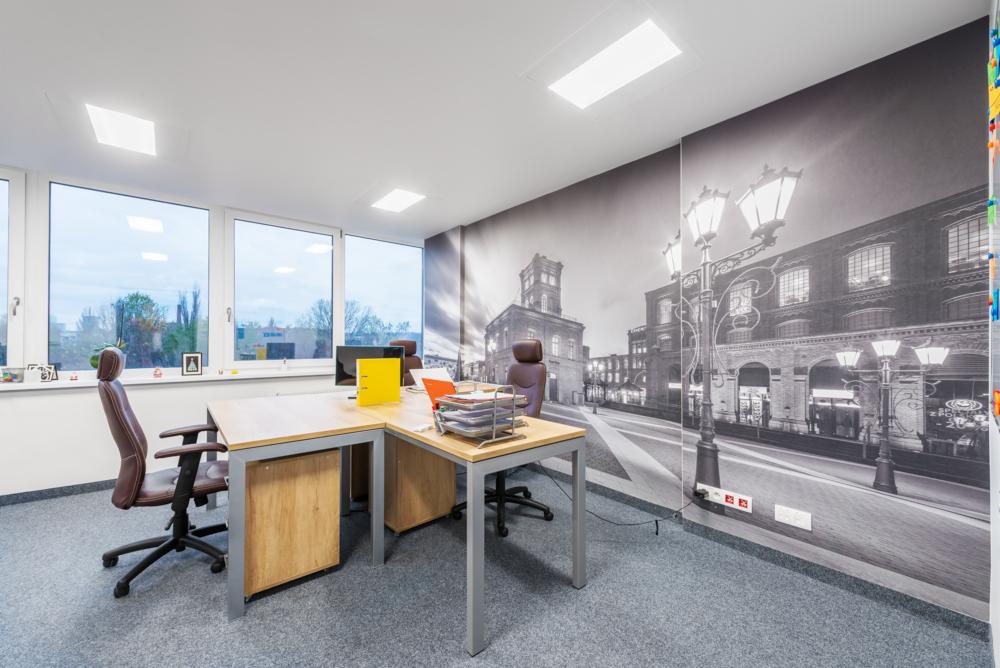 Jasne biuro z dużym oknem, biurkami, krzesłami i fototapetą przedstawiającą starą fabrykę z latarniami.