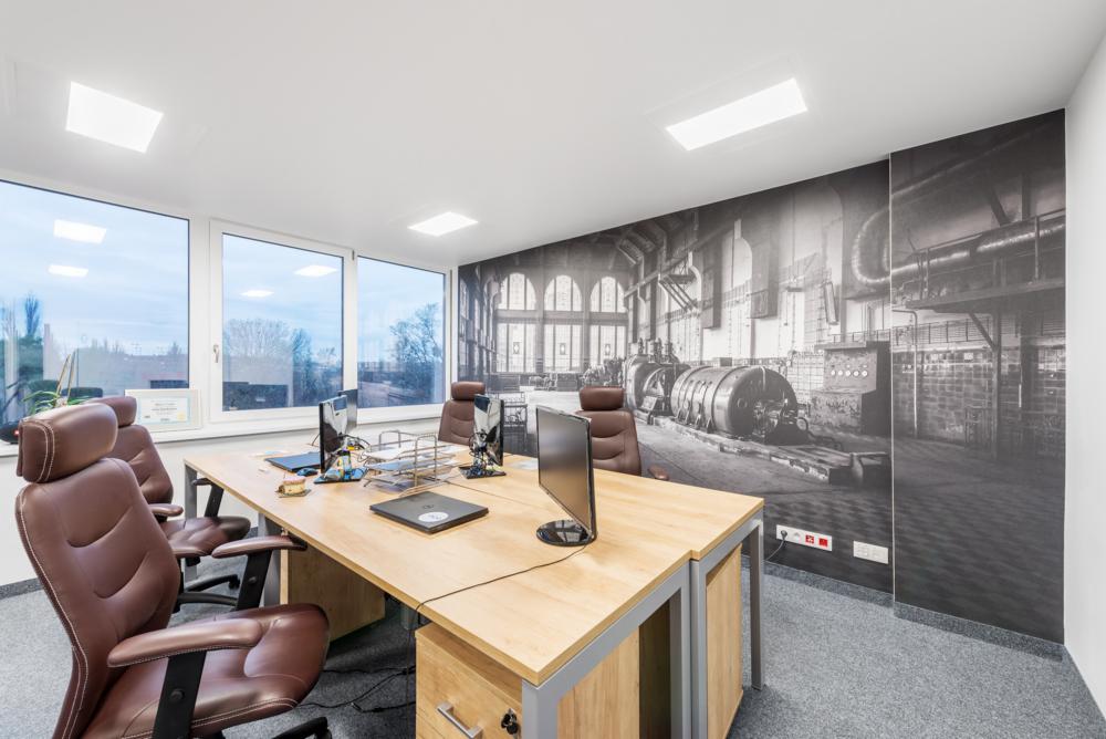 Jasne biuro z drewnianym biurkiem, skórzanymi krzesłami i fototapetą przedstawiającą wnętrze hali fabrycznej.