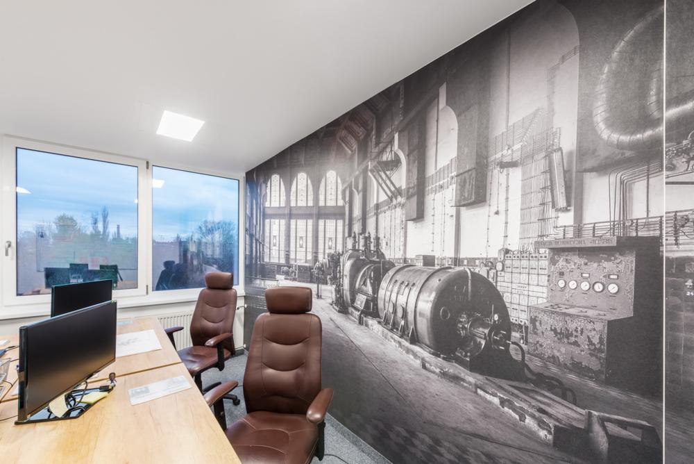 Nowoczesne biuro z fototapetą przedstawiającą wnętrze starej elektrowni, brązowe skórzane fotele obrotowe, drewniane biurko z monitorami i oknami z widokiem na zewnątrz.
