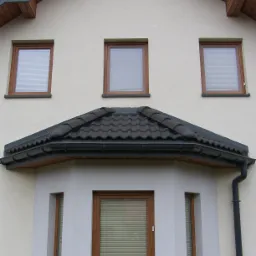 Elewacja domu z oknami PCV w drewnianej okleinie i żaluzjami poziomymi, widoczny fragment dachu z ciemną dachówką i rynna spustowa.