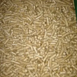 Jasne, drewniane granulki pelletu ułożone gęsto, z widocznym zielonym fragmentem krawędzi pojemnika w prawym górnym rogu.