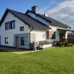 Ocieplony dom jednorodzinny z jasną elewacją, ciemną dachówką i klimatyzatorem LG na zewnątrz, otoczony zielonym trawnikiem w słoneczny dzień.