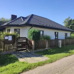 Jednopiętrowy dom z białą elewacją i ciemną dachówką w Miedźnie, widoczny z zewnątrz, z brązowym ogrodzeniem i zadbanym trawnikiem.