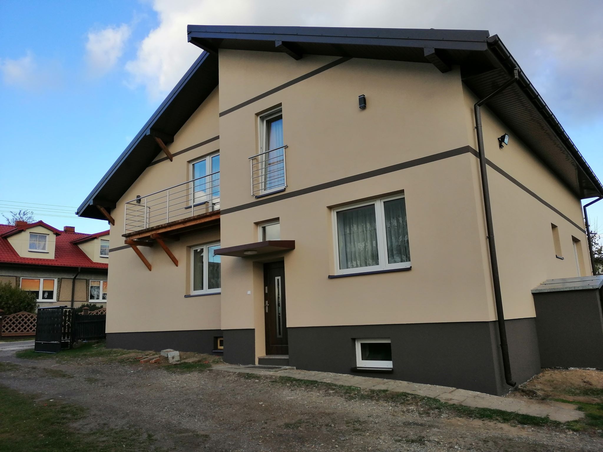 Dwukondygnacyjny dom z beżową elewacją, ciemnobrązowymi akcentami i balkonem z metalową balustradą, widoczny z bliska na tle pochmurnego nieba.