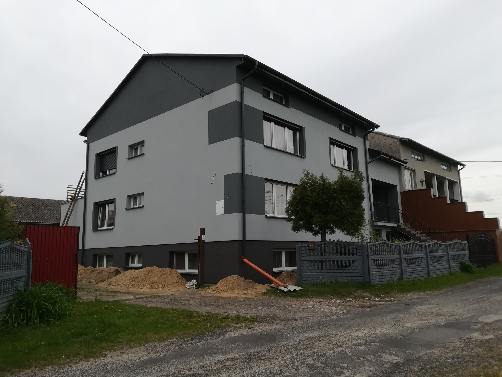 Dwukondygnacyjny dom z szarą elewacją w dwóch odcieniach, z widocznym rusztowaniem i stertami piasku przed budynkiem, sugerującymi prace budowlane.