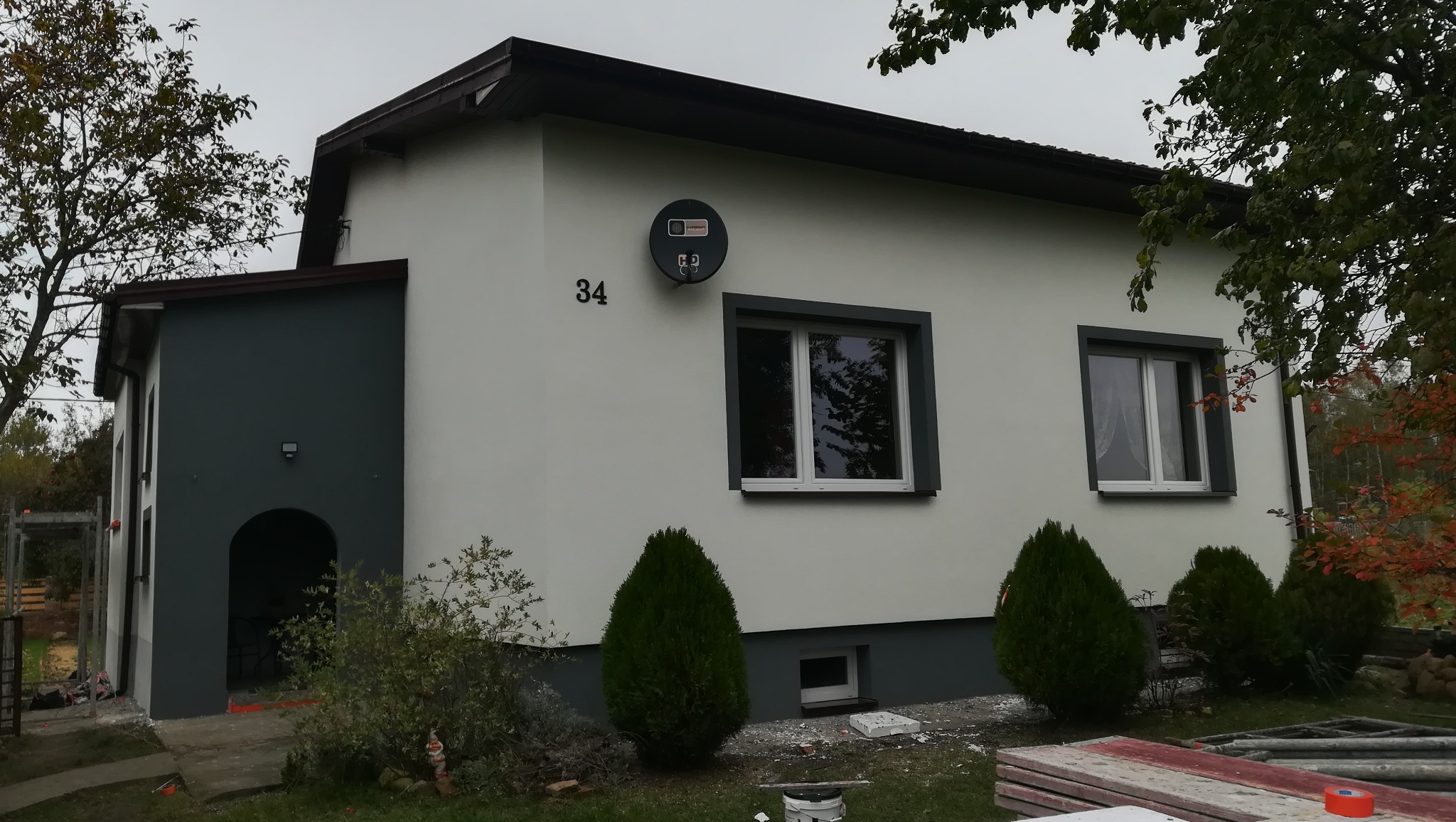 Dwukondygnacyjny dom z jasną elewacją i ciemnymi obramowaniami okien, numerem 34 na ścianie oraz satelitą, otoczony zielenią i fragmentem rusztowania.