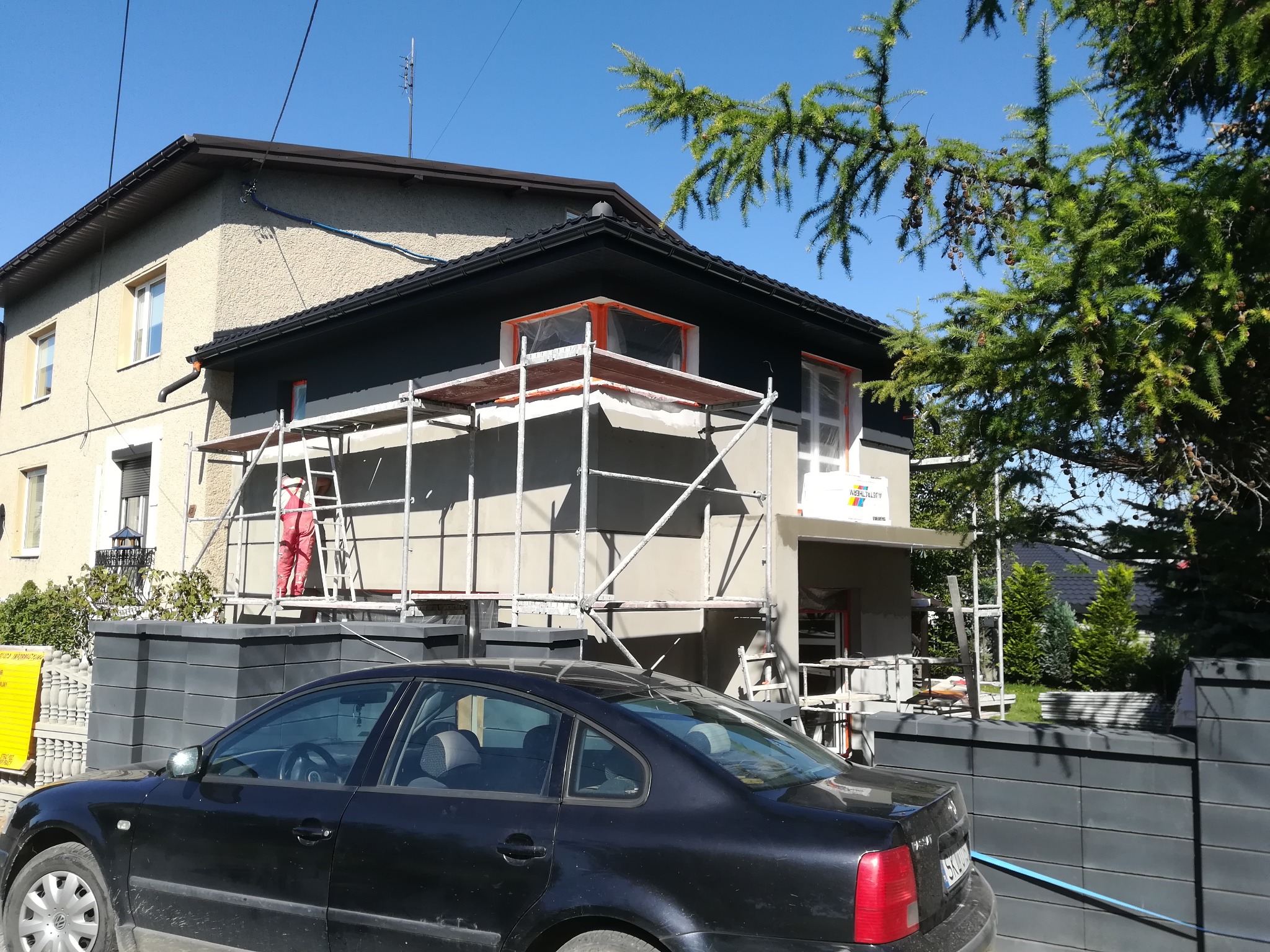 Dom w trakcie ocieplania elewacji, widoczne rusztowania, pracownik w czerwonym kombinezonie oraz częściowo otynkowana ściana w kolorze szarym, na pierwszym planie czarny samochód.