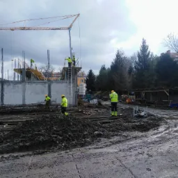 Budowa budynku żelbetowego w trakcie: robotnicy w odblaskowych kamizelkach montują zbrojenie, w tle wysoki dźwig budowlany, ponure niebo.