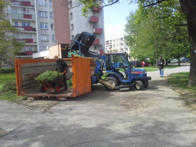 Pracownik z taczką pełną zielonych odpadów wrzuca je do pomarańczowego kontenera, obok stoi niebieski traktor z podnośnikiem, w tle blok mieszkalny i osoba przechodząca chodnikiem.