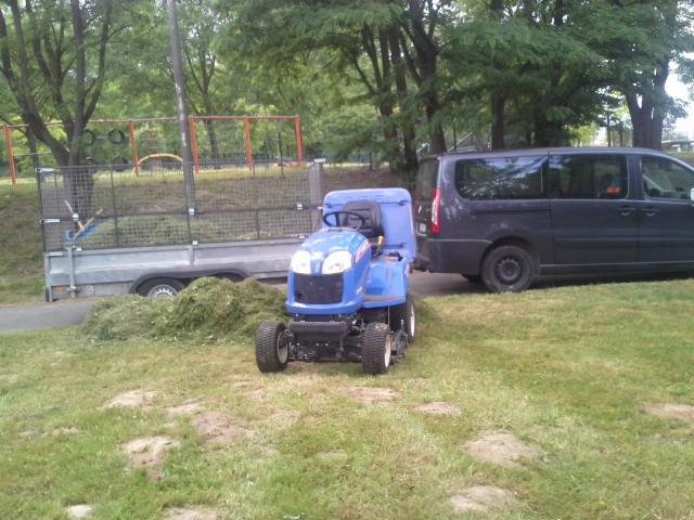 Niebieska kosiarka samojezdna z przyczepą pełną skoszonej trawy, zaparkowana obok ciemnego vana na trawniku w parku.