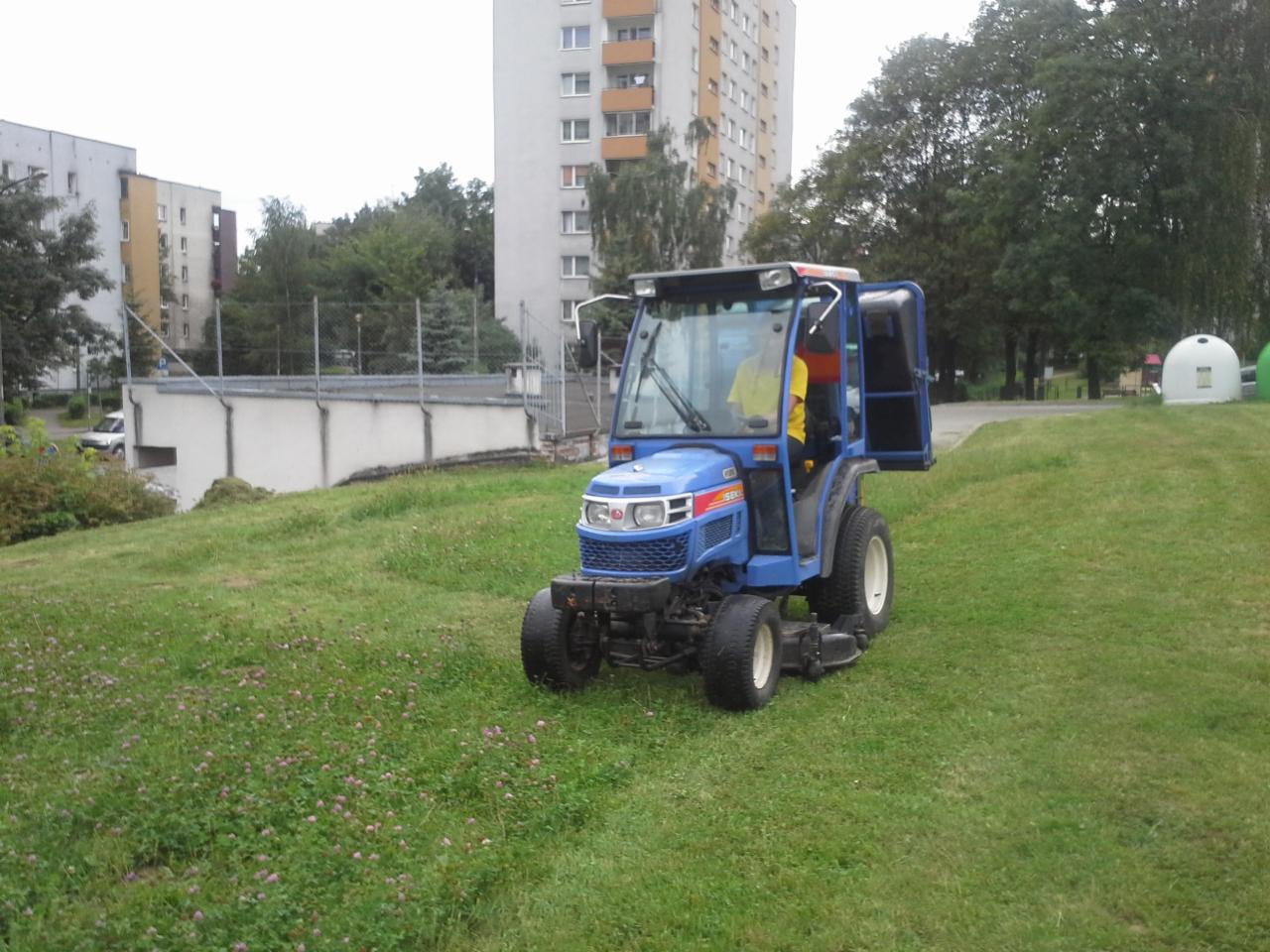 Niebieski traktor z otwartymi drzwiami kabiny na trawniku z kwitnącą koniczyną, w tle bloki mieszkalne i drzewa.
