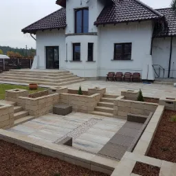 Nowoczesny taras z kostki brukowej w odcieniach beżu i szarości, otoczony podwyższonymi rabatami z roślinnością, z domu wychodzą szerokie schody, w tle biały dom z ciemną dachówką.